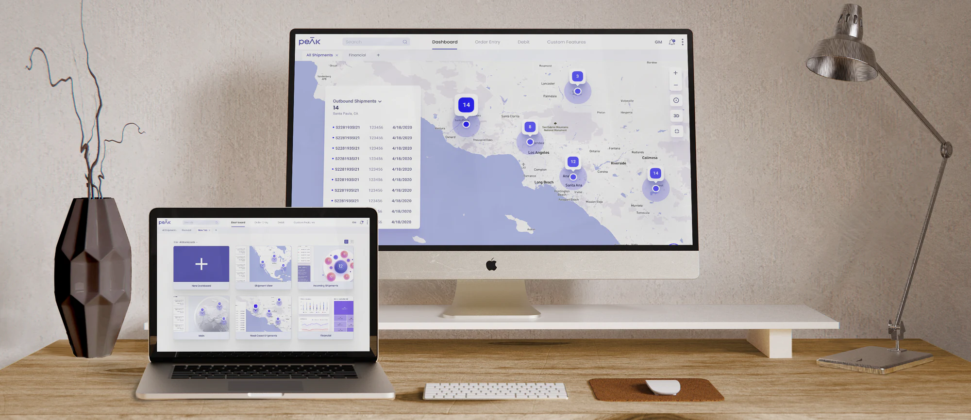 The PEAK dashboard on an iMac and a MacBook on a wooden desk: the 2D shipment map on the monitor, the widget dashboard on the laptop
